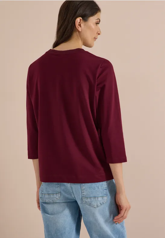 Frau von hinten mit dunkelrotem 3/4-Arm-Shirt und blauen Jeans vor beigem Hintergrund.