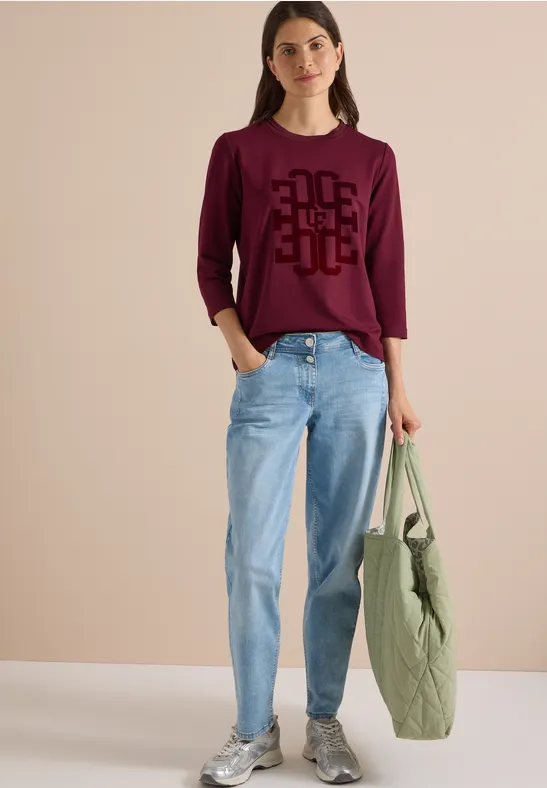 Frau in burgunderrotem Pullover mit schwarzem Muster, hellen Jeans, grauen Sneakers, trägt grünen Stepp-Shopper.
