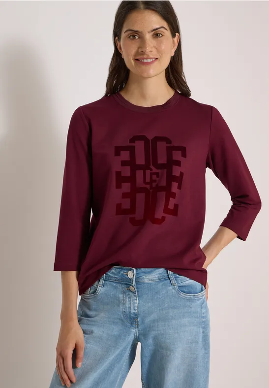 Frau trägt weinrotes 3/4-Arm-Shirt mit geometrischem Print und blaue Jeans.
