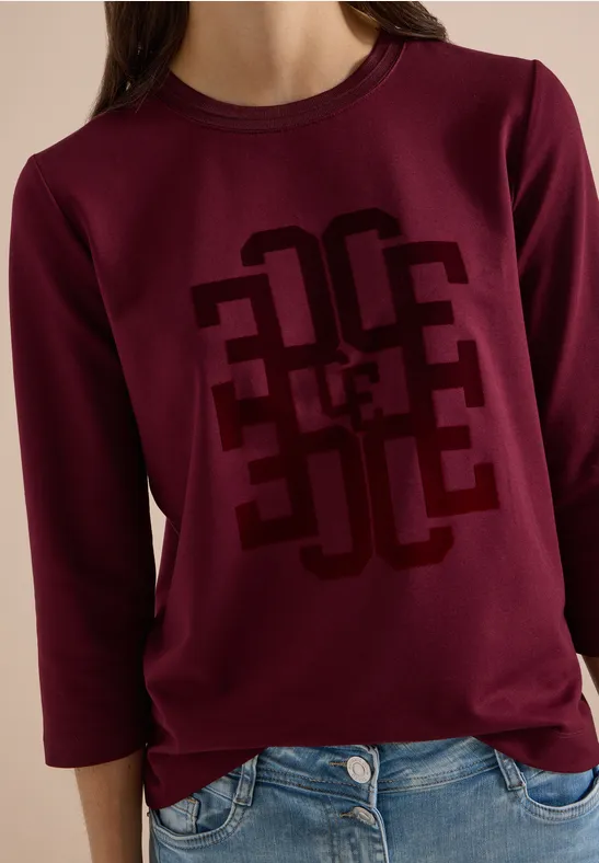 Frau trägt burgunderrotes Sweatshirt mit geometrischem Print und 3/4-Ärmeln, dazu blaue Jeans.