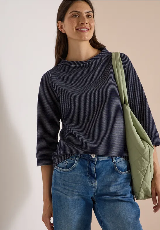 Junge Frau in Jeans und dunkelblauem Pullover trägt eine grüne Umhängetasche und lächelt.