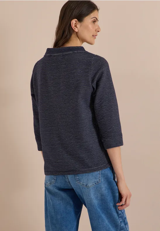 Frau von hinten trägt dunkelblauen Pullover mit 3/4-Ärmeln und Jeans vor hellem Hintergrund.