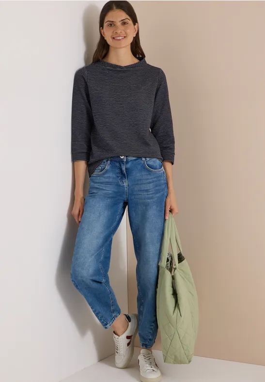 Frau in dunkelgrauem Langarmshirt, blauen Jeans und weißen Sneakern, hält eine hellgrüne Tasche.