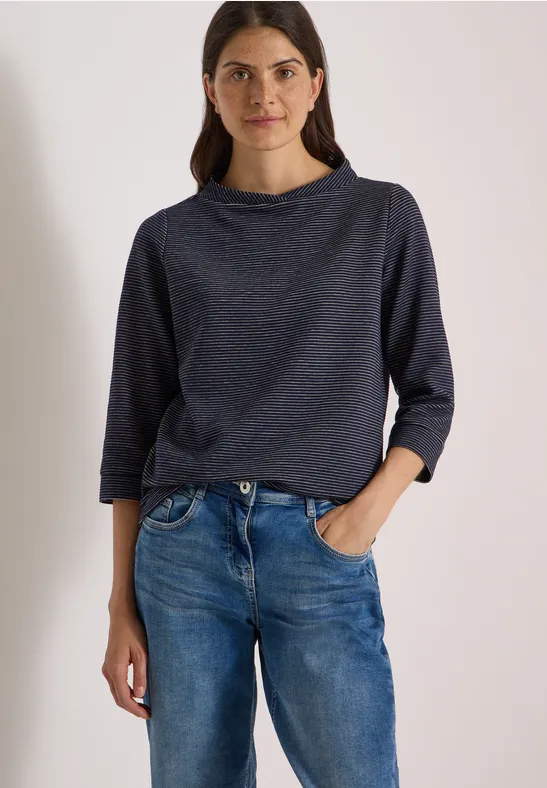 Frau steht lässig in Jeans und dunkelblauem Langarmshirt, eine Hand locker in der Hosentasche.