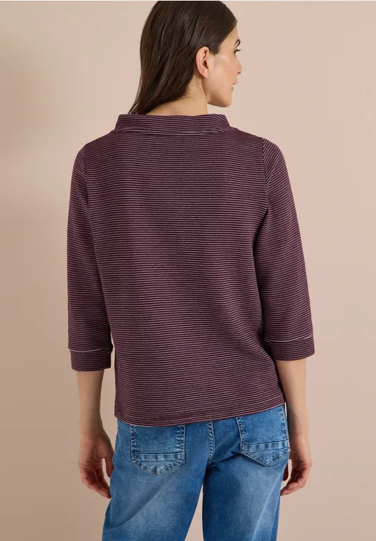 Frau steht mit dem Rücken zur Kamera, trägt lila Pullover und Jeans.