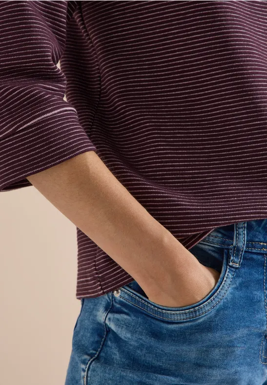 Person mit bordeaux-weiß gestreiftem Langarmshirt und blauen Jeans, Hand in der Hosentasche.