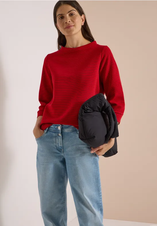 Frau in rotem Pullover, Jeans und mit schwarzer Jacke vor neutralem Hintergrund.