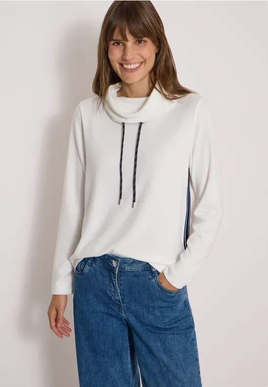 Lächelnde Frau in weißem Rollkragenpullover mit schwarzen Kordeln und Jeans vor hellem Hintergrund.