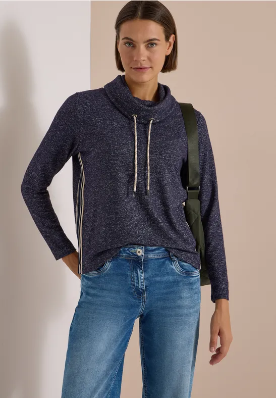 Femme en pull bleu marine col boule à cordons et jean, posant décontractée devant un mur beige.
