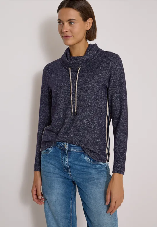 Femme portant un pull bleu foncé à cordon et un jean, debout devant un mur clair.