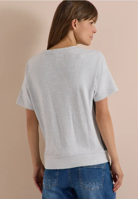 Rückansicht einer Frau mit kurzärmeligem, hellgrauem Stricktop und blauen Jeans vor beigem Hintergrund.