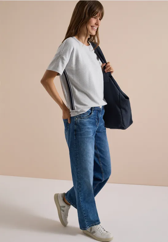 Frau in weißem T-Shirt und weiten Jeans trägt eine blaue Tasche und weiße Sneaker vor hellem Hintergrund