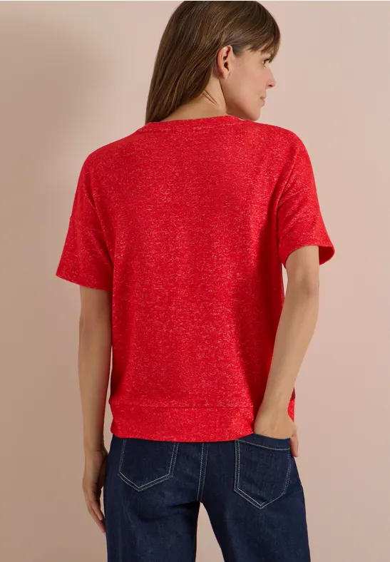 Frau von hinten in rotem Kurzarmshirt und blauen Jeans vor beigem Hintergrund.