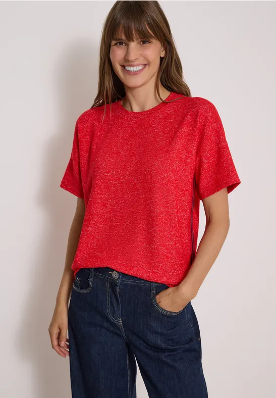 Lächelnde Frau in rotem Kurzarmshirt und blauer Jeans, eine Hand in der Hosentasche.