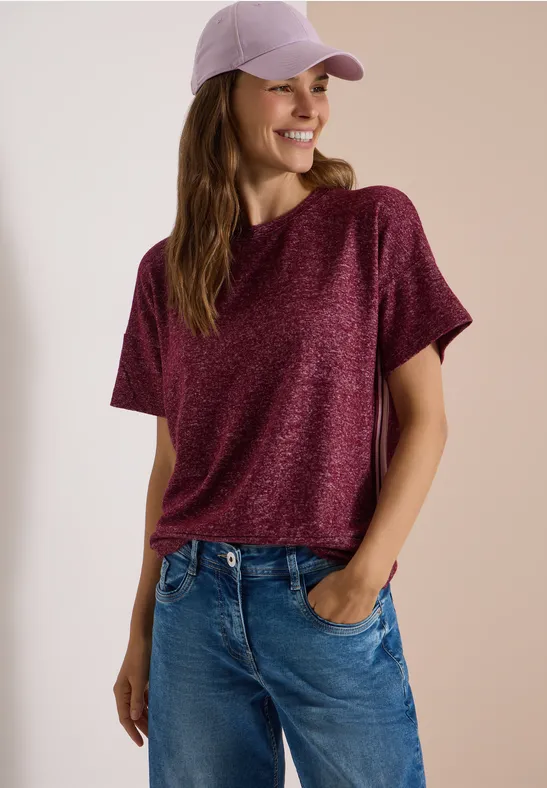 Frau trägt lila Cap, lockeres bordeauxfarbenes Shirt und Jeans, lächelt und hat Hände in den Taschen.