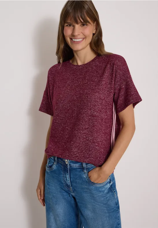 Frau in rotem T-Shirt und blauer Jeans lächelt vor neutralem Hintergrund.