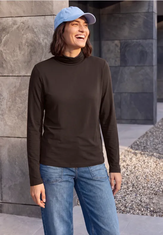 Frau im schwarzen Langarmshirt, Jeans und blauer Kappe lächelt vor einer steinernen Wand im Freien.