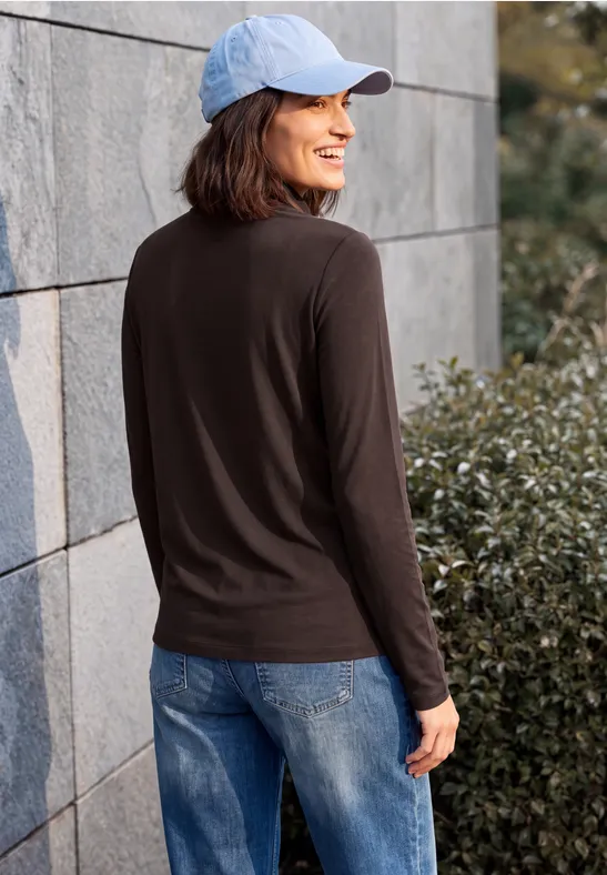 Frau im hellblauen Cap, schwarzem Langarmshirt und Jeans steht lächelnd an einer Mauer vor Büschen.
