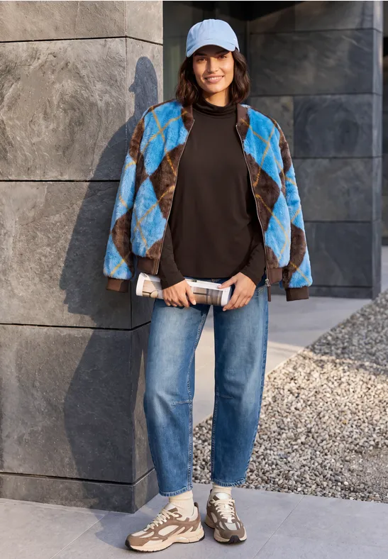 Frau mit Cap, blackem Shirt, blau gemusterter Jacke, Jeans und Sneakern lehnt an grauer Steinmauer