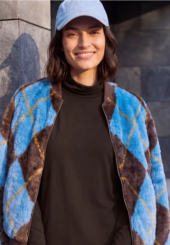 Frau mit blau-brauner Rautenjacke, schwarzem Rollkragenpullover und heller Cap lächelt vor grauer Wand.