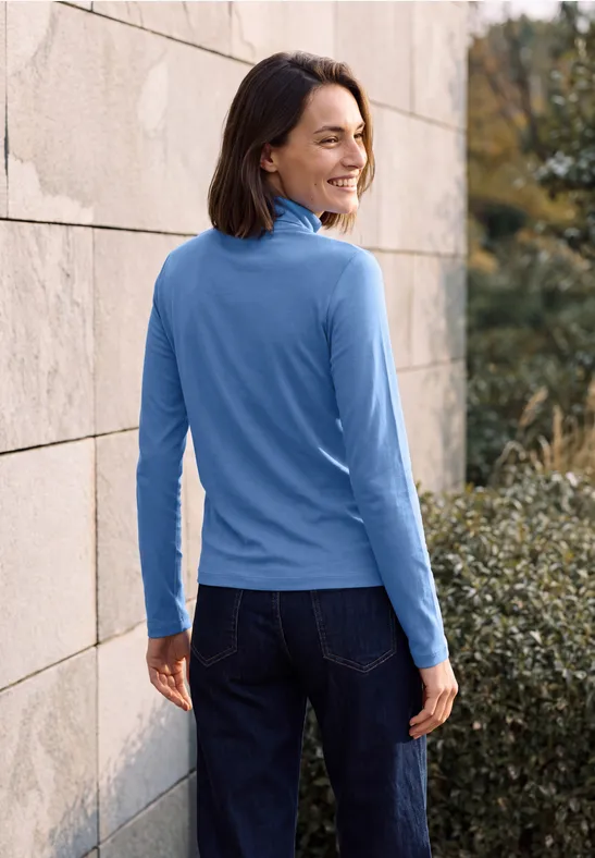 Frau in blauem Langarmshirt und Jeans lehnt an einer Steinwand und lächelt über die Schulter.