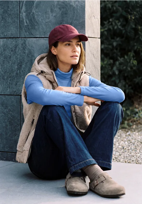 Eine junge Frau im lässigen Outfit sitzt auf dem Boden, lehnt an einer Steinmauer und schaut nach rechts.
