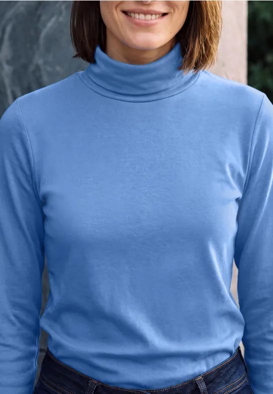 Eine Frau trägt einen hellblauen Rollkragenpullover und Jeans, leicht lächelnd.