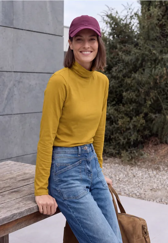 Eine Frau mit gelbem Rollkragenpullover, Jeans und pinker Mütze lehnt lächelnd an einer Holzbank.