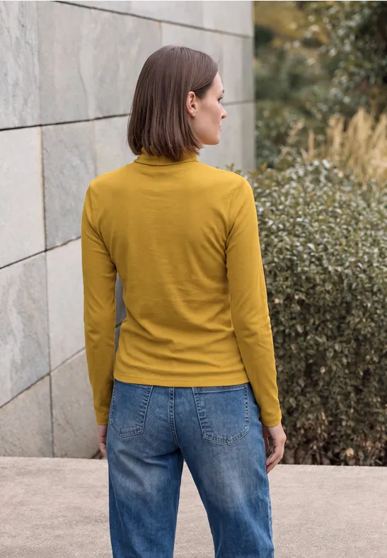 Frau in gelbem Langarmshirt und Jeans steht im Freien vor einer Steinwand und Büschen.