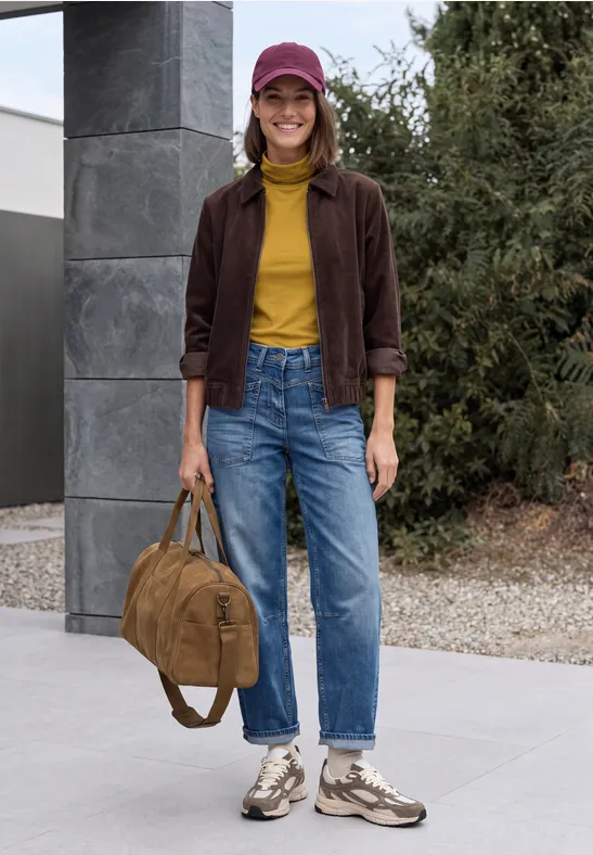 Junge Frau in gelbem Rollkragenpullover, brauner Jacke, Jeans, Sneakers und Mütze hält einen Rucksack.