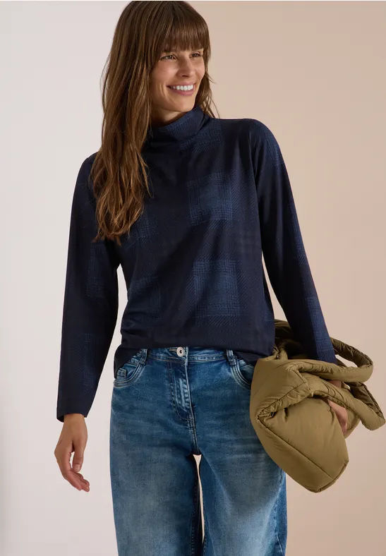 Frau in dunkelblauem Rollkragenpullover und Jeans hält eine olivgrüne, geknotete Tasche und lächelt.