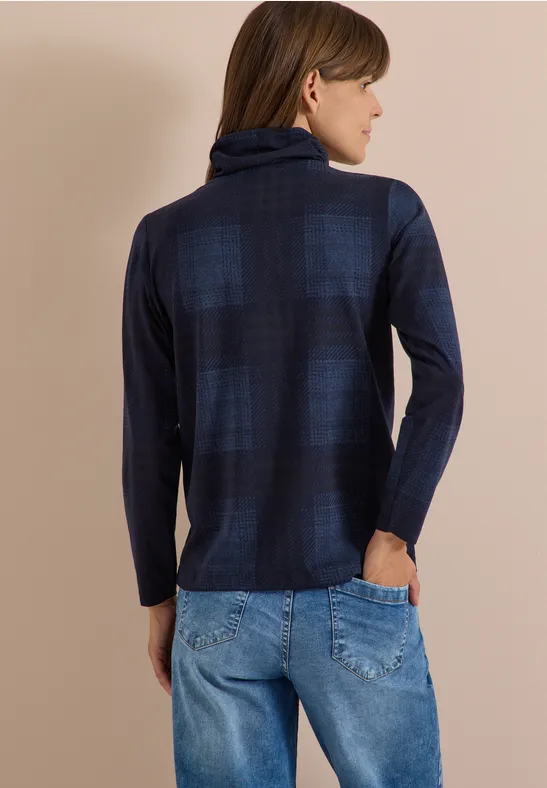 Frau steht seitlich, trägt dunkelblauen karrierten Rollkragenpullover und blaue Jeans.