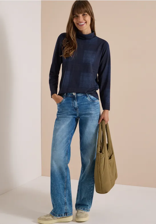 Frau in dunkelblauem Rollkragenpullover, weiten Jeans und olivgrüner Tasche vor neutralem Hintergrund.