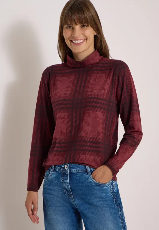 Eine Frau trägt ein bordeauxrotes Karopullover mit Rollkragen und blaue Jeans, lächelnd.