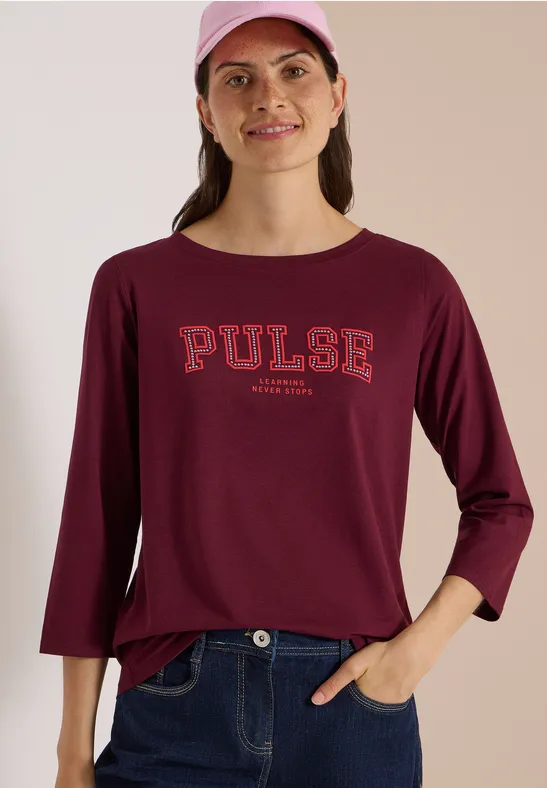 Femme en tee-shirt bordeaux à manches longues 'PULSE', jean et casquette rose, souriante.