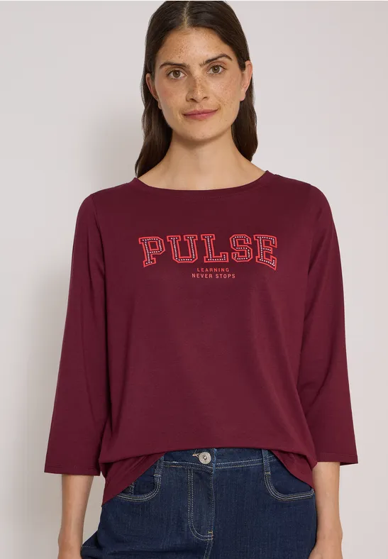 Femme portant un t-shirt bordeaux avec l'inscription 'PULSE' et un jean foncé.