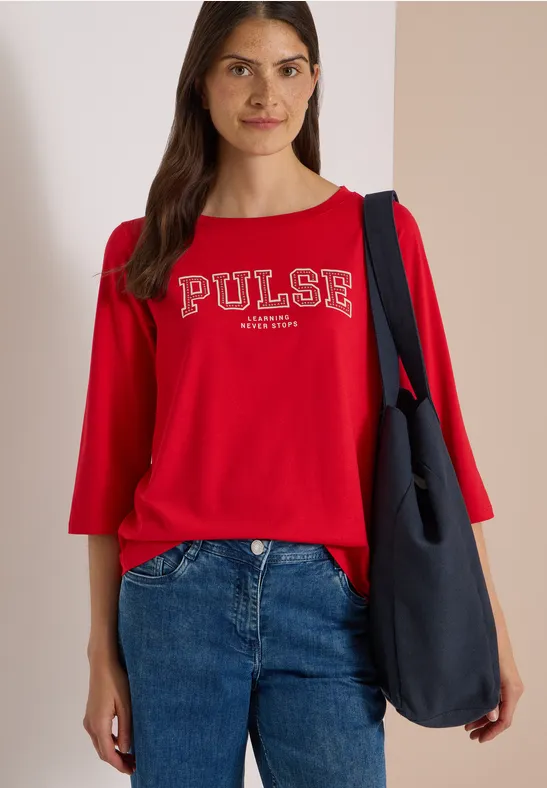 Femme en pull rouge 'PULSE', jean bleu et sac bleu foncé, devant un mur clair.
