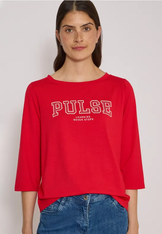 Jeune femme devant fond clair porte un t-shirt rouge 'PULSE' et un jean bleu.
