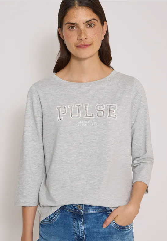 Jeune femme en t-shirt gris 'PULSE' et jeans, souriante devant un fond clair, une main dans la poche.