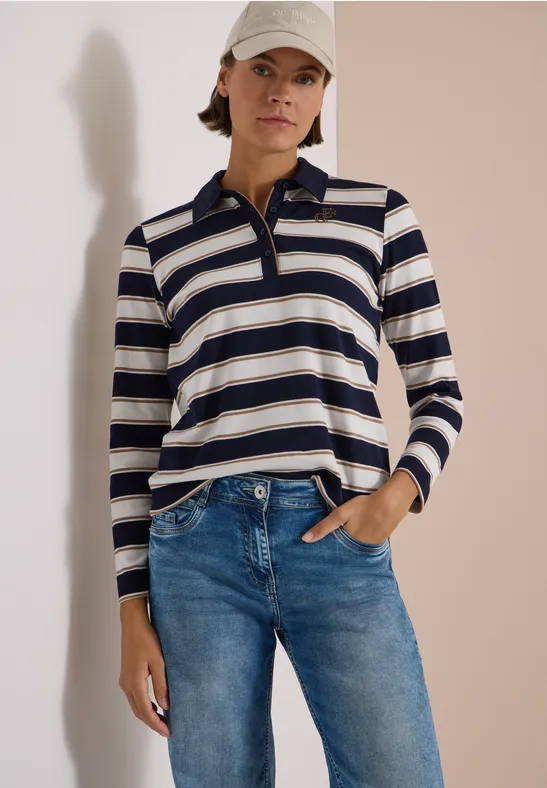 Femme porte un polo rayé à manches longues, un jean et une casquette, la main dans la poche.