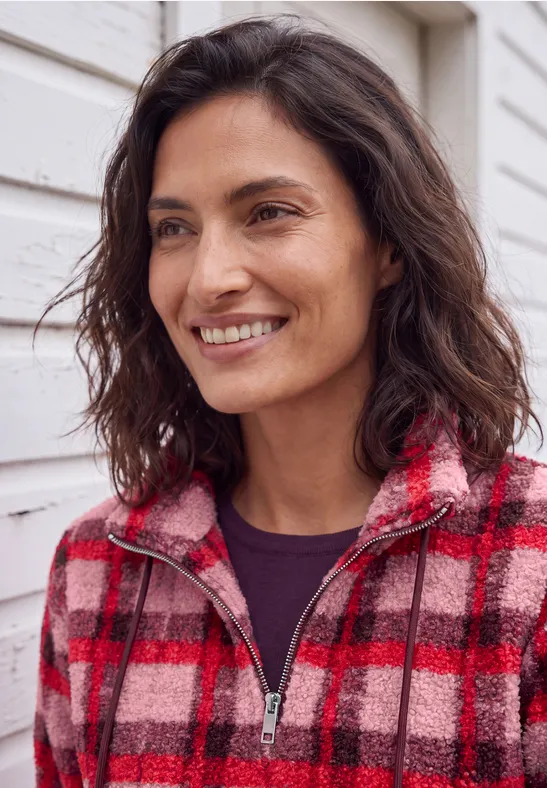 Eine lächelnde Frau in einem rot-karierten Fleecejacke vor einer weißen Hauswand.