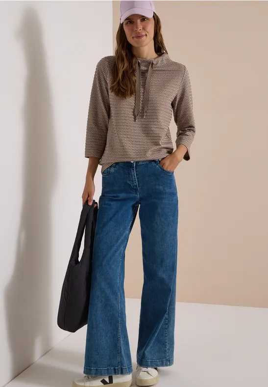 Frau mit gestreiftem Kapuzenpullover, rosa Cap, weiten Jeans und großer schwarzer Tasche vor hellem Hintergrund.