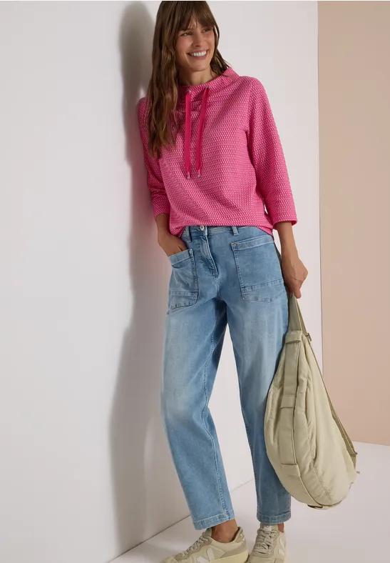 Junge Frau lehnt lächelnd an einer Wand, trägt rosa Kapuzenpullover, weite Jeans und eine große Beuteltasche.