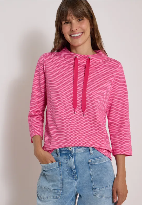 Frau trägt pinken, gemusterten Hoodie mit Dreiviertelärmeln und lockere blaue Jeans, lächelnd.