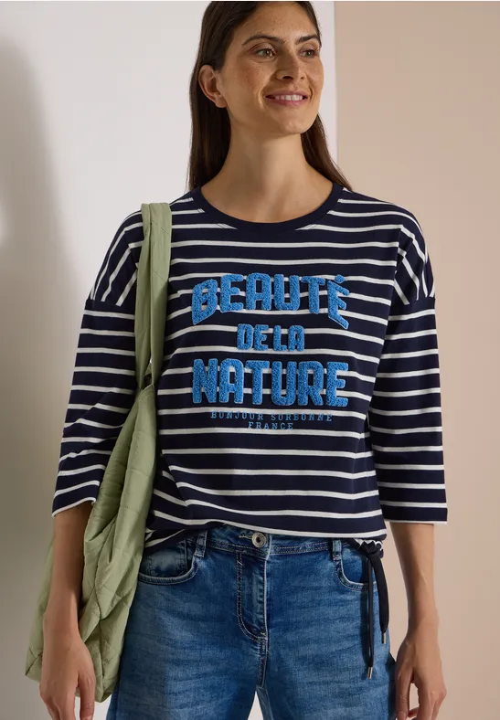 Frau in gestreiftem Shirt mit „Beauty of Nature“-Aufdruck, Jeans und grüner Jacke über der Schulter.