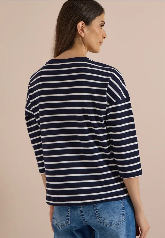Frau von hinten in gestreiftem Marineblau-Weiß-Shirt und Jeans vor beigem Hintergrund.