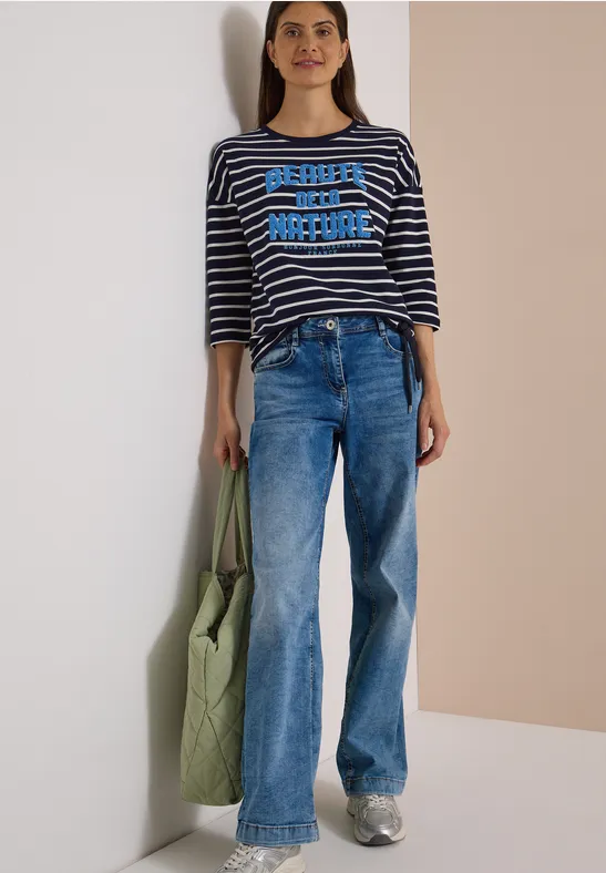 Eine Frau in blauer Jeans, gestreiftem Shirt und grüner Tasche lehnt entspannt an einer hellen Wand.