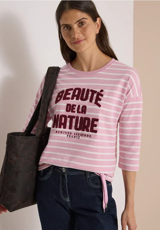 Frau trägt rosa-weiß gestreiftes Shirt mit Aufschrift 'Beauté de la nature', Jeans und schwarze Tasche.