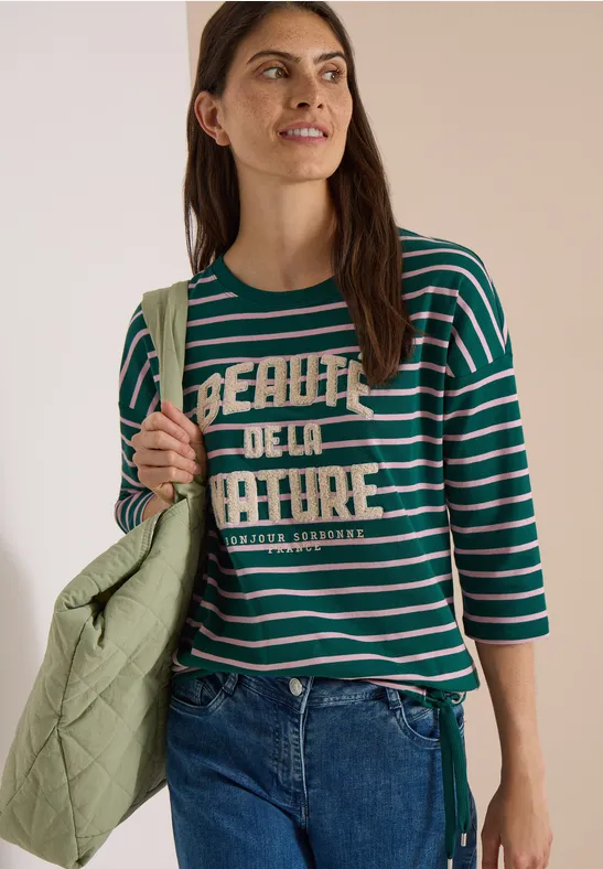Frau trägt grün-weiß gestreiftes Shirt mit Schriftzug, Jeans und gesteppte Tasche.