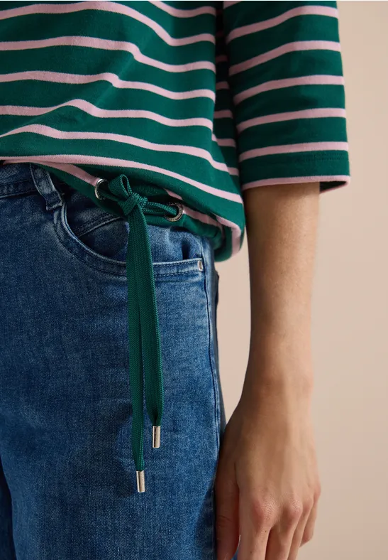 Person trägt grün-weiß gestreiftes Oberteil und blaue Jeans mit grünem Taillengürtel, Hand sichtbar